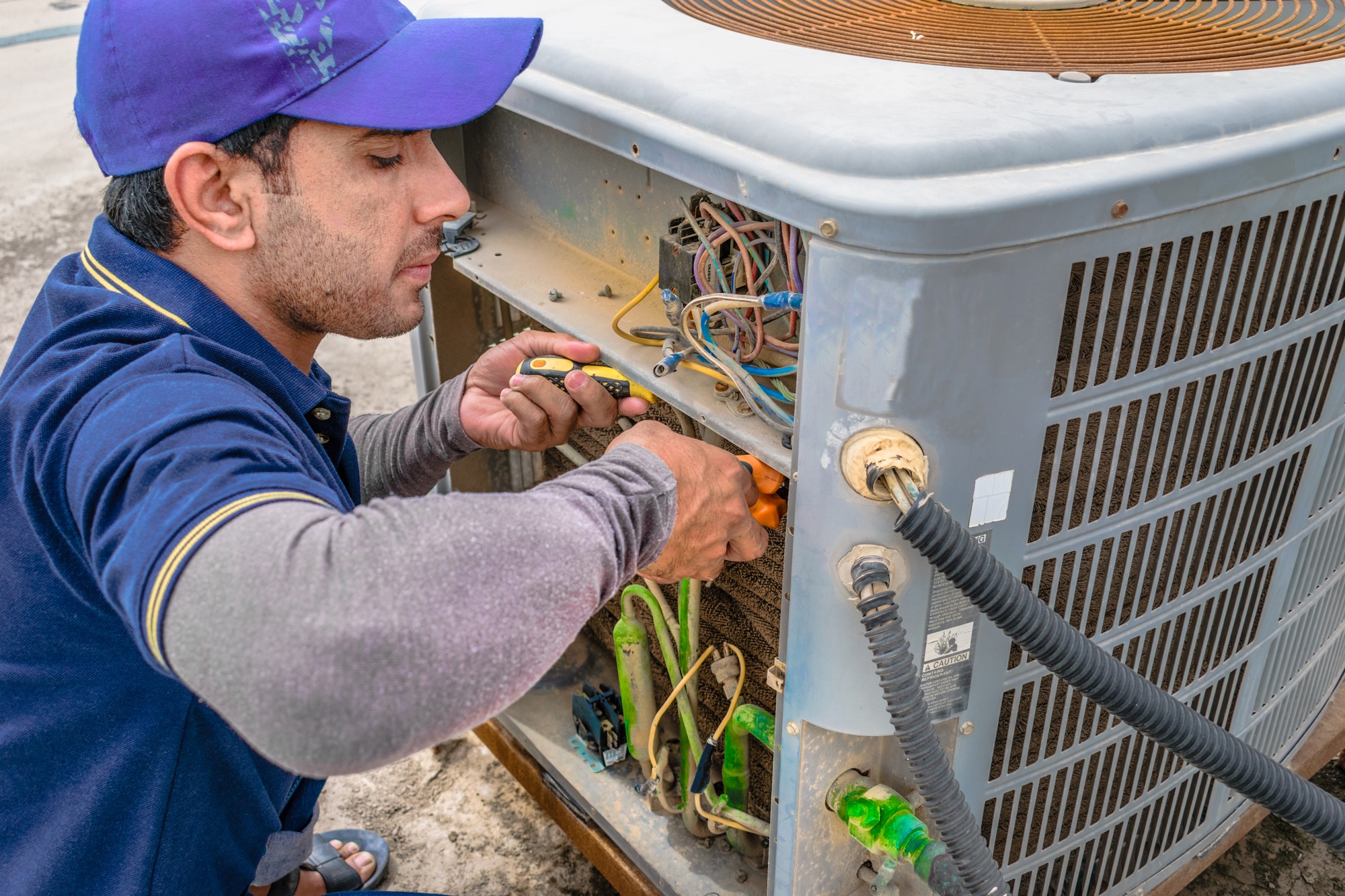 Technician repairs an outdoor air conditioning unit, focusing on electrical connections and components. The image highlights HVAC maintenance work.