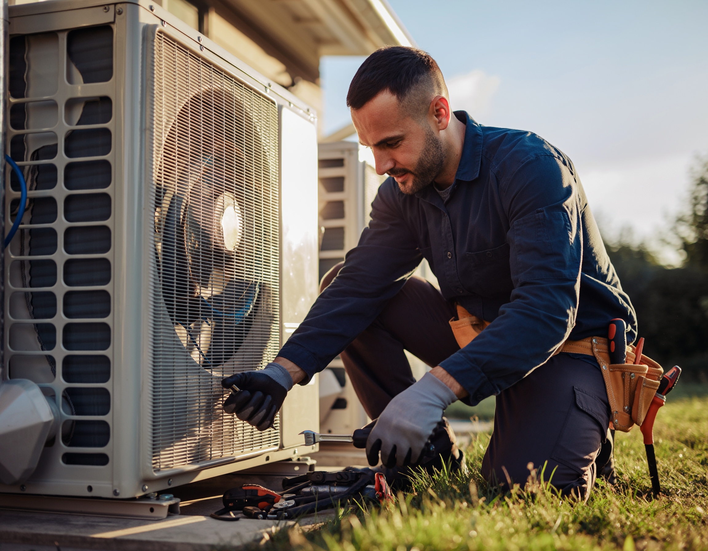 Technician servicing an outdoor air conditioning unit, using tools and wearing gloves, focused on maintenance in a grassy area at sunset.
