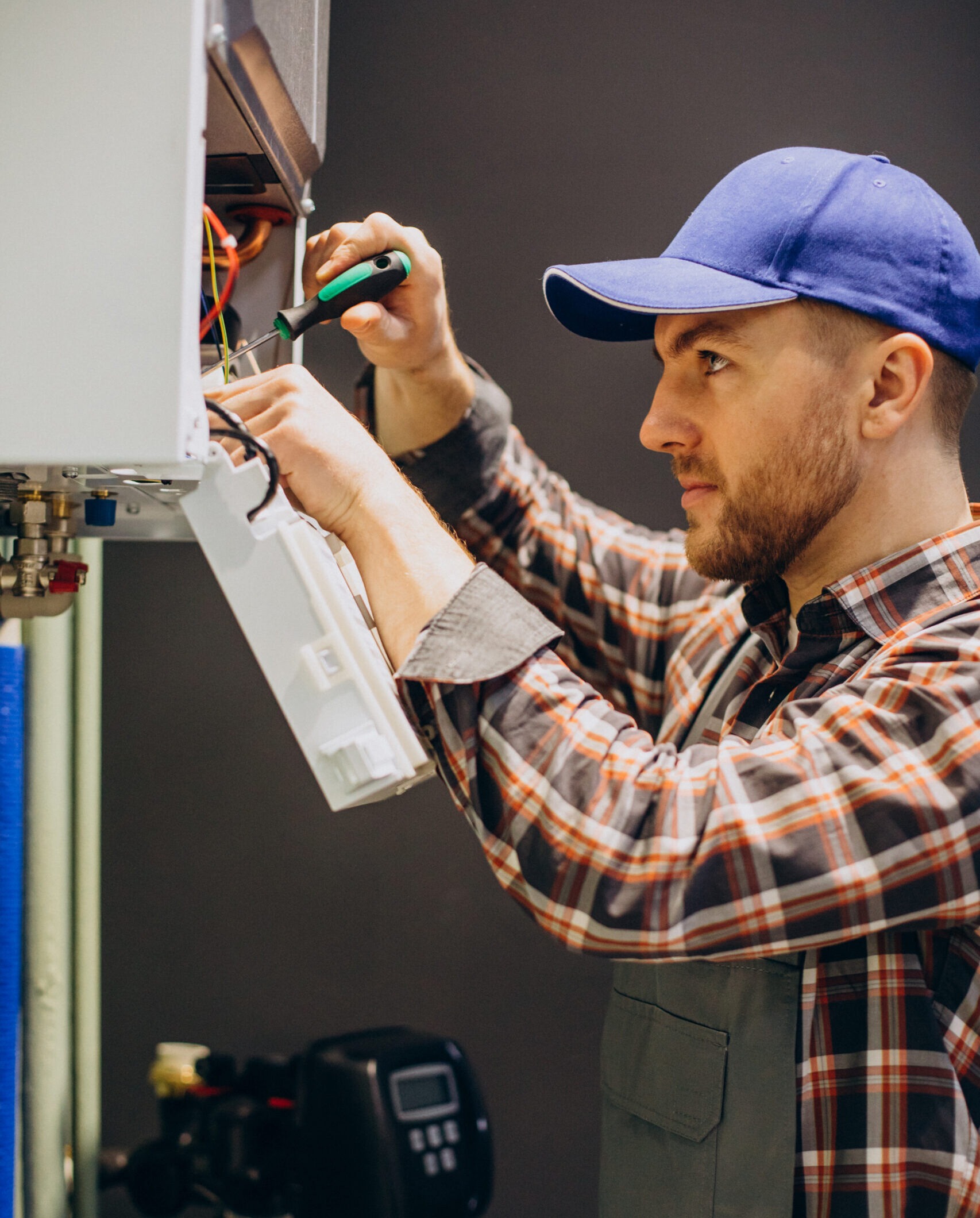 Technician in a plaid shirt and blue cap repairs a heating unit, using a screwdriver to connect wires, emphasizing hands-on maintenance work.